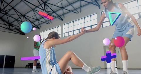 Female basketball teammate helping up seated player on indoor court with neon 3d overlays