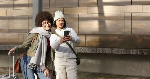 Young women checking smartphone while traveling with rolling suitcase against wood-panel wall
