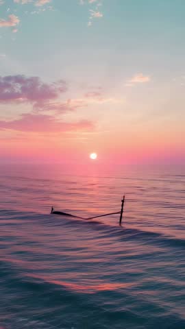 Vertical footage of boat drifting on calm ocean at pastel sunset with mast silhouette