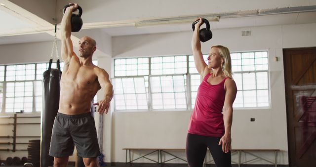 Pairs training together in gym lifting kettlebells high above their heads with focused expressions. Ideal for fitness blogs, exercise guides, training programs, gym advertisements, and health promotion materials.
