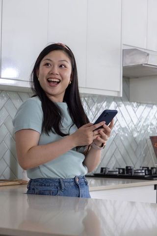 Young asian woman smiling while holding smartphone in modern bright kitchen lifestyle home