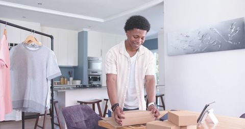 Young entrepreneur packing ecommerce orders at kitchen table with clothing rack and tablet
