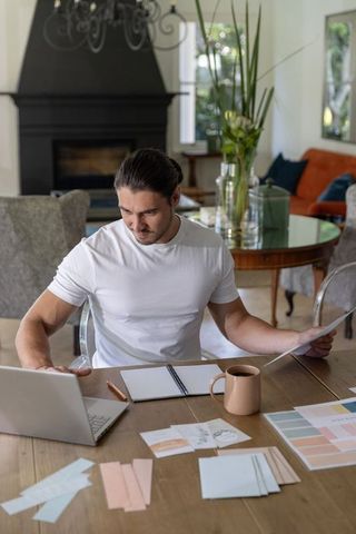 Designer working from home reviewing color swatches at sunlit dining table with laptop