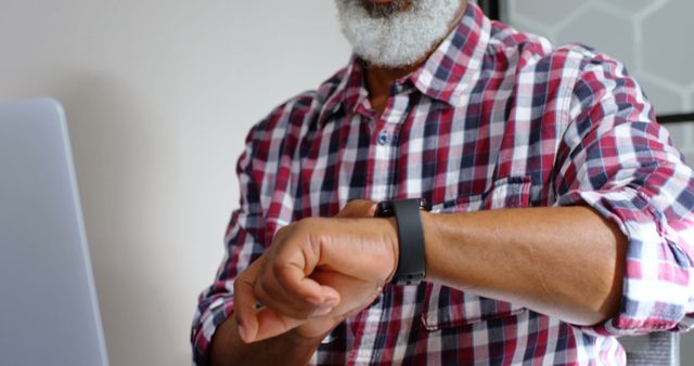 Senior man with beard checking smartwatch while sitting in modern office wearing a plaid shirt. Useful for depicting technology use by older adults, productivity in office environments, and modern workplace settings. Ideal for illustrating themes such as time management, work-life balance, and advancements in wearable technology.