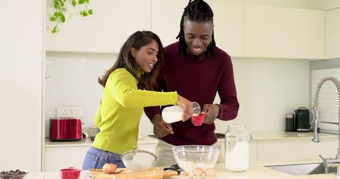 Smiling diverse couple baking together in modern minimalist kitchen pouring milk into bowl