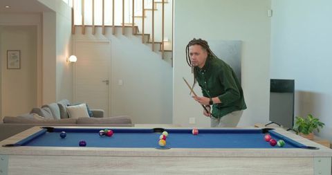 African-american man leaning and aiming cue on blue pool table in modern living room
