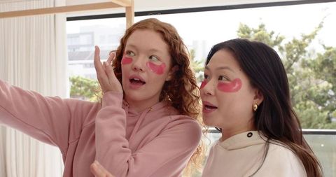 Surprised diverse friends wearing pink under-eye patches in hoodies on bright balcony
