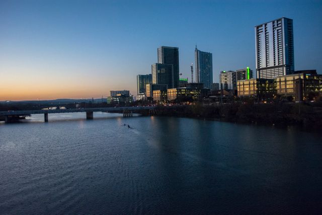 This photo captures the serene riverfront of a modern city at dusk. The high-rise buildings reflect in the still water, providing a contrast to the vibrant evening sky. Ideal for use in real estate, urban planning, travel, and tourism content that highlights modern architecture and city living.