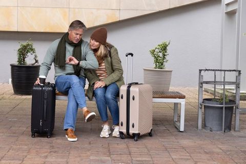 Mature couple waiting on bench with luggage checking watch and holding coffee outdoors
