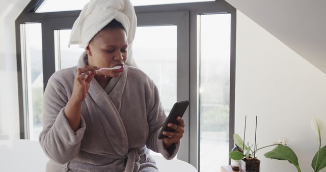 This image shows a woman brushing her teeth while looking at her smartphone in a modern bathroom. She is wearing a bathrobe and has a towel wrapped around her head. The scenery suggests a casual morning routine. Ideal for use in content related to personal hygiene, morning routines, multitasking, or self-care.