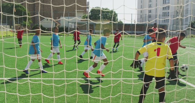 Group of children wearing uniforms playing soccer on a green field through a netting view. The boys are divided into two teams, one in red uniforms and one in blue, actively engaging in the game. Ideal for promoting youth sports, teamwork, and healthy lifestyle activities. Suitable for use in education materials, sports advertisements, and community program promotions.