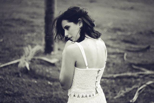 Young woman wearing a lace dress standing in a natural outdoor area, seen from behind in a pensive pose. The image is in black and white, adding a vintage and nostalgic feel. Ideal for use in blogs, articles, or advertisements focusing on themes of introspection, romance, and nature. Perfect for fashion editorials, creative projects, or print media emphasizing a dreamy atmosphere.