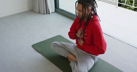 African american man practicing breathwork and meditation on yoga mat by sunlit balcony door
