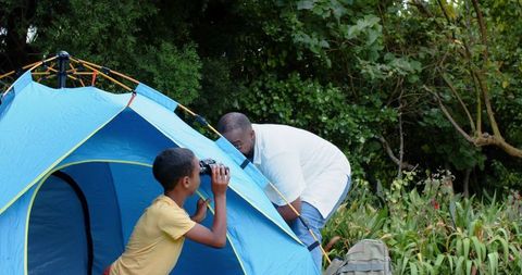 African american father and son camping in backyard at blue tent, exploring with binoculars