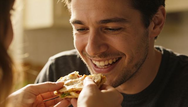 Smiling man being fed flatbread by partner in cozy kitchen, intimate breakfast moment