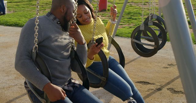 Couple sitting on swings in a park, smiling and looking at a mobile phone, enjoying each other's company. This image can be used for relationship or leisure time concepts, advertisements for parks or outdoor activities, or illustrating bonding and affection between couples.
