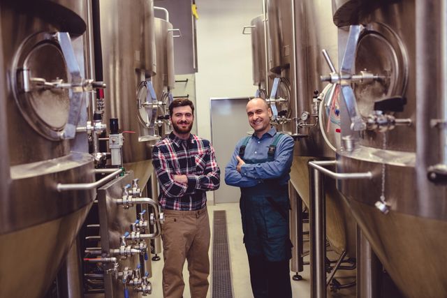 Two confident brewery workers standing amidst large fermentation tanks, showcasing teamwork and professionalism in an industrial setting. Ideal for use in articles about the brewing industry, teamwork in manufacturing, or professional profiles in industrial environments.