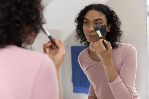 Non-binary adult applying charcoal face mask at mirror wearing pink top and jewelry