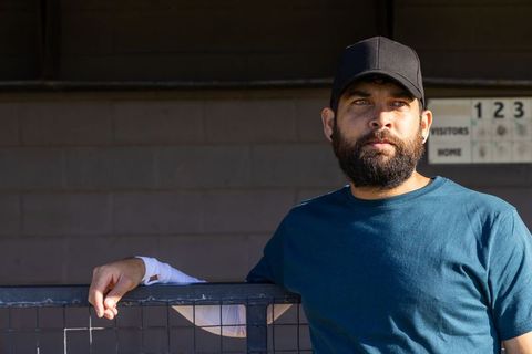 Sunlit coach leaning on dugout fence wearing cap and teal t-shirt, thoughtful look