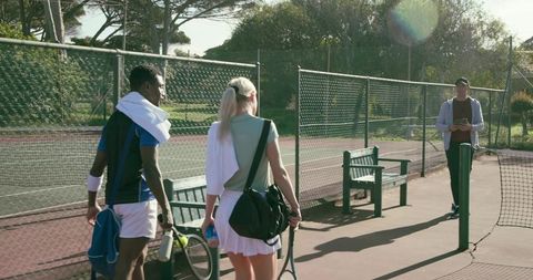 Diverse tennis pair walking toward coach on outdoor court during sunny training session