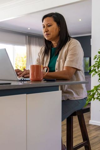 Mature asian woman working from home at kitchen island typing on laptop with coffee mug