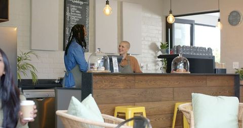 Diverse Baristas Chatting and Smiling Behind Wood Counter in Sunlit Cozy Cafe with Pastries