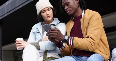Asian woman and african american man sharing smartphone on urban bench with coffee