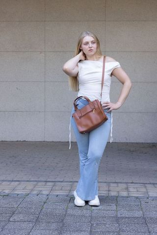 Woman standing on city sidewalk wearing off-shoulder top, flared jeans, brown leather bag