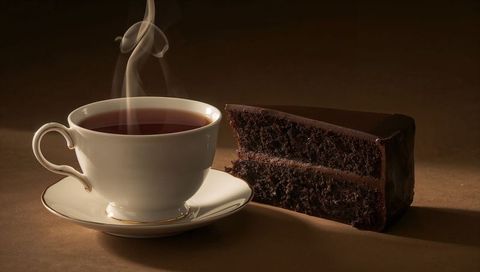 Steaming porcelain teacup with rich chocolate cake slice on warm brown backdrop in soft light