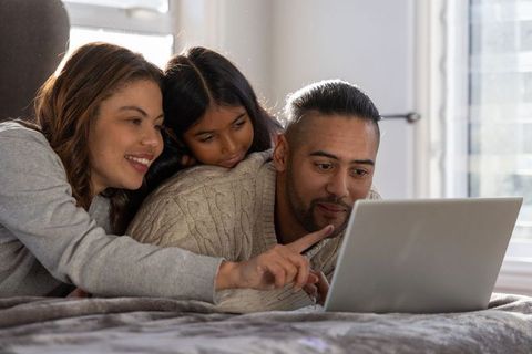 Family browsing laptop together on bed, parents and daughter sharing cozy screen time