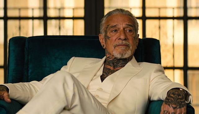 Tattooed senior gentleman reclining in cream suit on teal velvet chair with backlit window