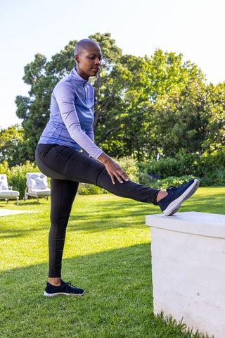 African woman stretching leg on low ledge in sunny garden practicing outdoor fitness