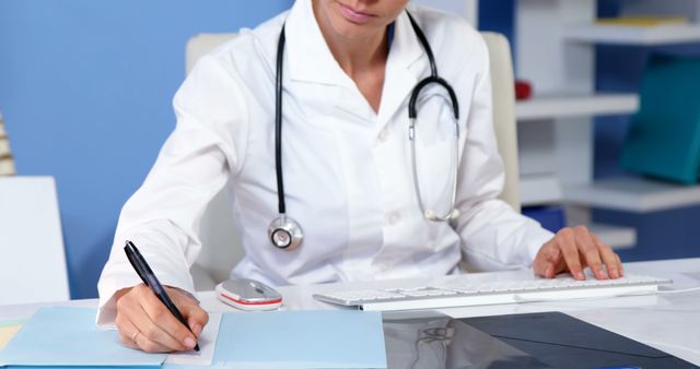 Healthcare professional wearing white coat and stethoscope, sitting at desk, writing medical notes while working on computer. Suitable for concepts related to medical documentation, healthcare industry, professional routine, doctor's office organization, and health management.