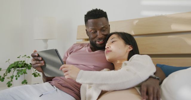 Happy diverse couple using tablet and lying in bedroom. Spending quality time at home concept.