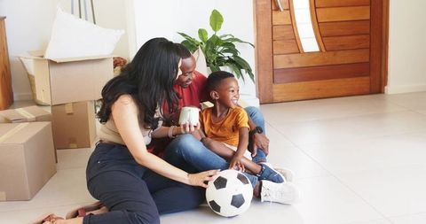 Joyful family settling into new home with moving boxes, parents and son enjoying soccer