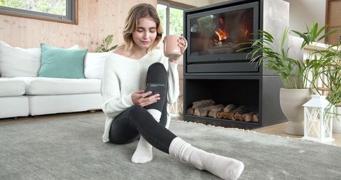 Woman Relaxed at Home Using Smartphone Near Cozy Fireplace