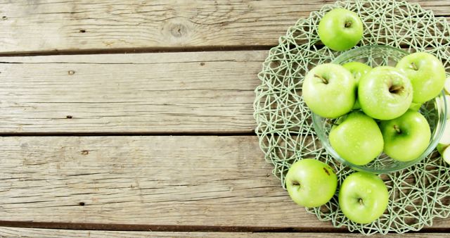 Green apples arranged in glass bowl with green woven mat, placed on rustic wooden table. Fresh and organic, suitable for promoting healthy eating, farm produce, autumn harvest themes, or natural kitchen decor. Great for food blogs, restaurant websites, and nutrition articles.