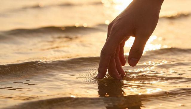 Hand touching calm shoreline water at sunset with golden ripples and soft reflection