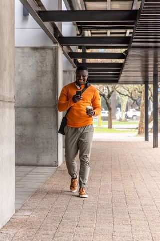 African american man walking with coffee and phone in orange sweater and headphones
