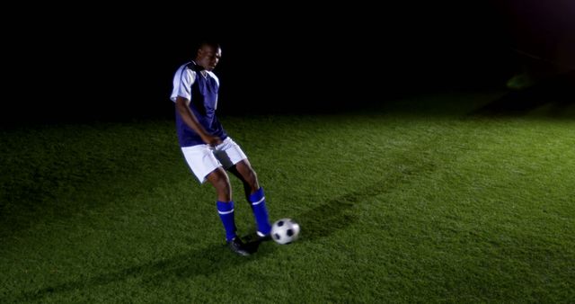 A young African American male athlete is practicing soccer on a grassy field at night, with copy space. His focused expression and dynamic posture capture the dedication required in sports training.