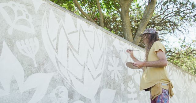 Artist painting intricate floral mural on outdoor wall surrounded by trees. Suitable for illustrating concepts of creativity, outdoor art projects, artistic expression, and public art installations.
