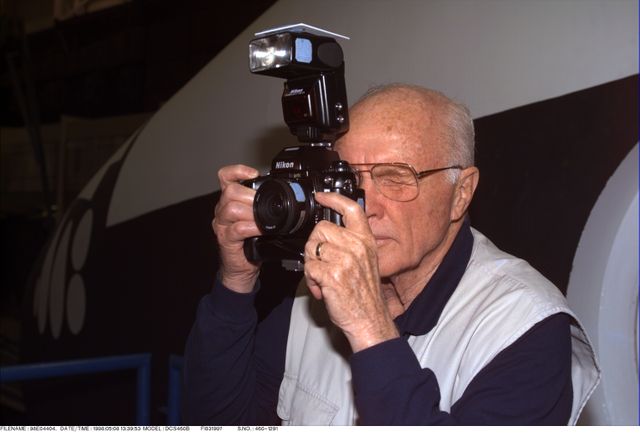 Senior man holding a 35mm camera with flash attachment in front of a NASA shuttle training facility. Meant to capture the dedication of space exploration and historical moments in NASA's history. Perfect for editorial use, historical retrospectives, and discussions on NASA training, astronaut preparedness, or commemorating milestone achievements.