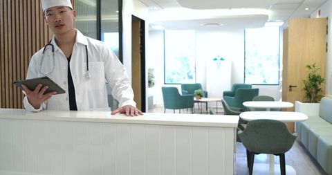 Doctor standing at modern clinic reception holding tablet and stethoscope, welcoming patients
