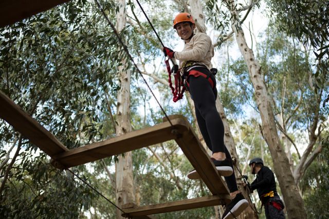 Woman crossing zip line in forest adventure park, smiling and wearing safety gear. Man in background also participating. Ideal for promoting outdoor activities, adventure parks, team-building events, and recreational sports.