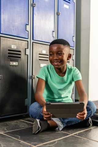 African american boy sitting by school lockers holding tablet smiling and learning