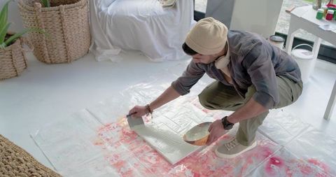 Indian artist wearing beanie crouching smoothing gesso on canvas in bright home studio