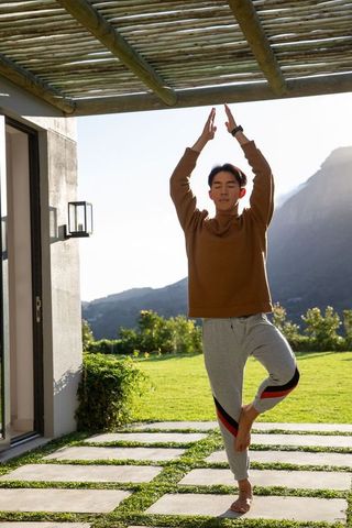 Young man practicing tree pose on sunny patio under wooden pergola with mountain view
