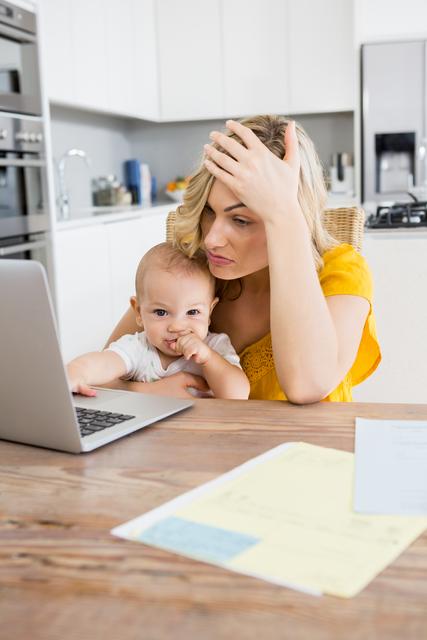 Tensed mother using laptop with her baby boy in kitchen at home