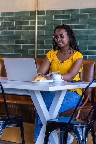 African american woman working on laptop in modern cafe with coffee, croissant and green tiles