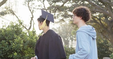 Sunlit campus friends celebrating graduation walking in gown and mortarboard with hoodie